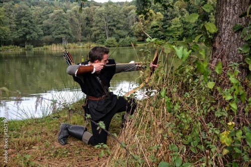 Mittelalterlicher Bogenschütze mit schwarzer Kapuze kniet seitlich am Boden vor einem See , zielt mit Pfeil und Bogen zur Seite 