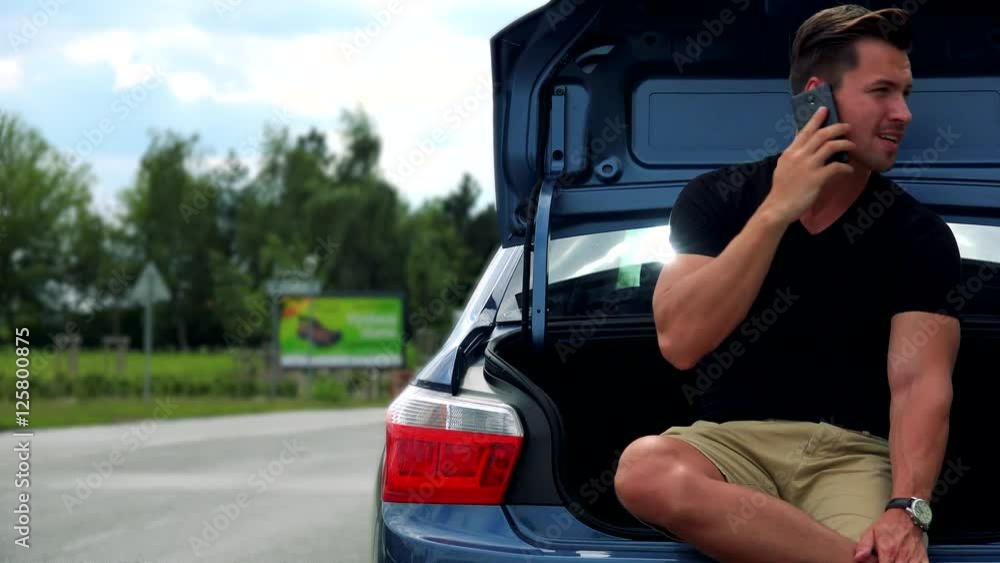 young handsome man sits in car trunk and phones with smartphone - road with cars young handsome man sits in car trunk and phones with smartphone - road with cars