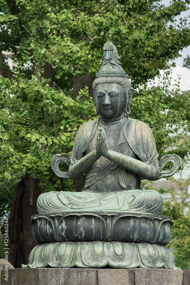 Tokyo, Japan - September 26, 2016: Closeup of bronze statue of Kannon Bosatsu, attendant of Amida Buddha, sitting on lotus pedestal in garden at Senso-ji Buddhist Temple. Green foliage. 