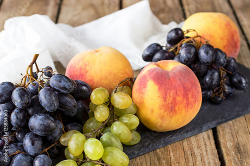A bunch of grapes and ripe peaches on a black slate board on a wooden table