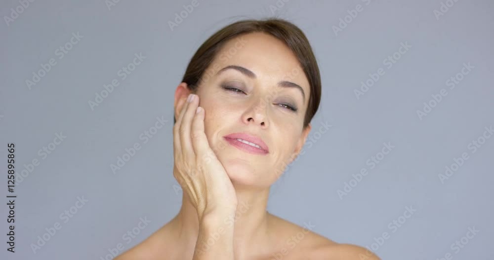Front view on cute single young adult woman with eyes closed and hands on cheeks over gray background with copy space Front view on cute single young adult woman with eyes closed and hands on cheeks over gray background with copy space