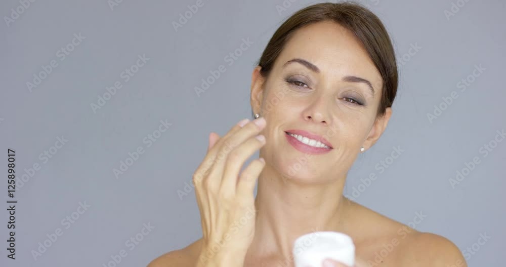 Gorgeous young woman applying face cream from a handheld jar in a beauty skincare makeup and glamour concept over grey with copy space Gorgeous young woman applying face cream from a handheld jar in a beauty skincare makeup and glamour concept over grey with copy space