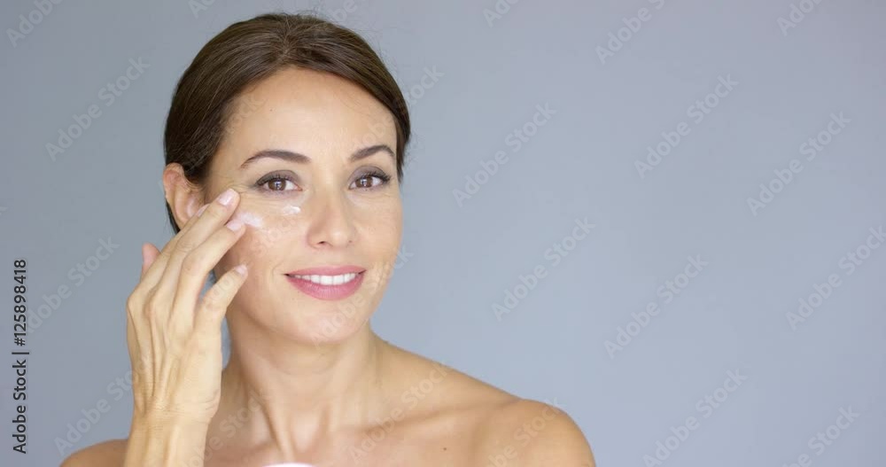 Beautiful young woman applying face cream to her cheek bone in a beauty skincare or cosmetics concept on grey with copy space Beautiful young woman applying face cream to her cheek bone in a beauty skincare or cosmetics concept on grey with copy space