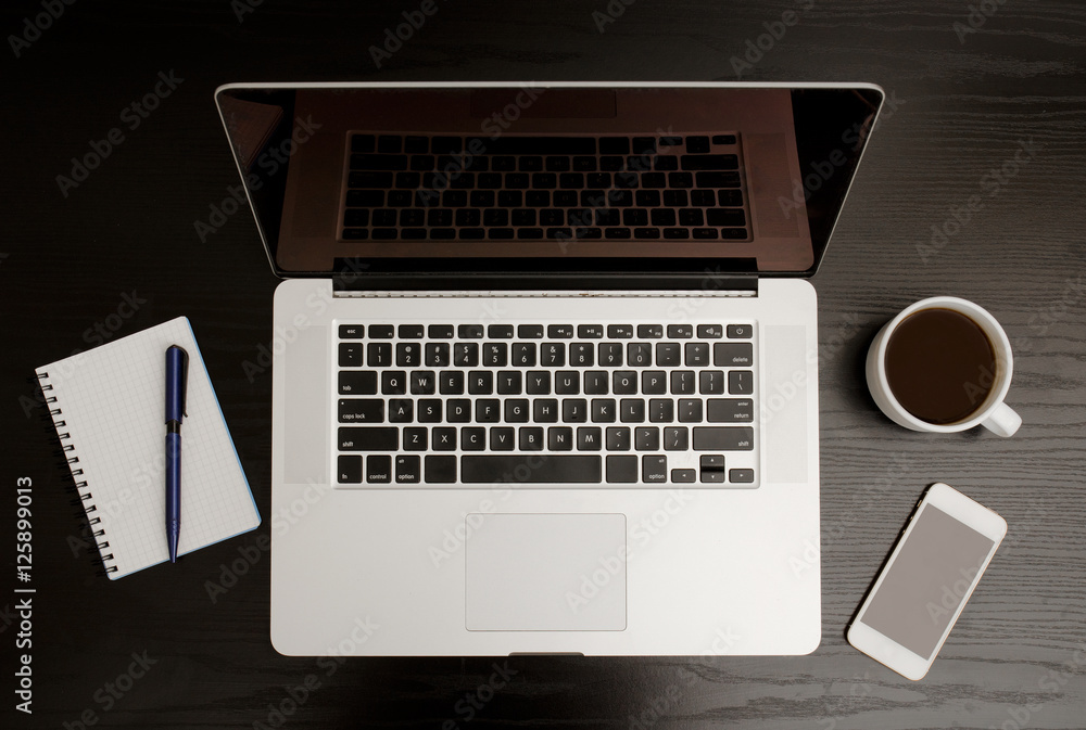 Top view of the open laptop, notebook with pen, smart phone and a mug of coffee on a black wooden table Top view of the open laptop, notebook with pen, smart phone and a mug of coffee on a black wooden table