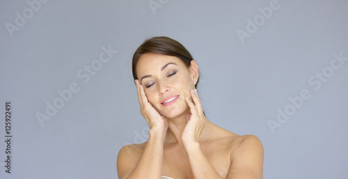 Front view on cute single young adult woman with eyes closed and hands on cheeks over gray background with copy space