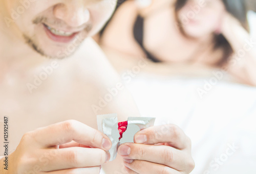 Man holding red condom with blurred background of a woman lie on a bed