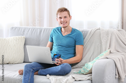 Young man with laptop sitting on sofa and making online shopping