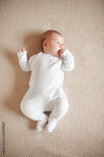 Pretty baby in a white pajamas lying on his back and holding  pen in  mouth