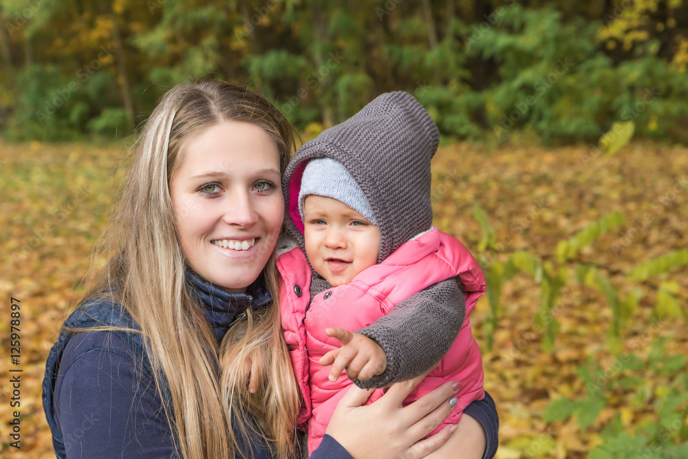 Smiling young mother with her baby daughter in arms is looking at the camera in beautiful autumn park on fall day. Smiling young mother with her baby daughter in arms is looking at the camera in beautiful autumn park on fall day.