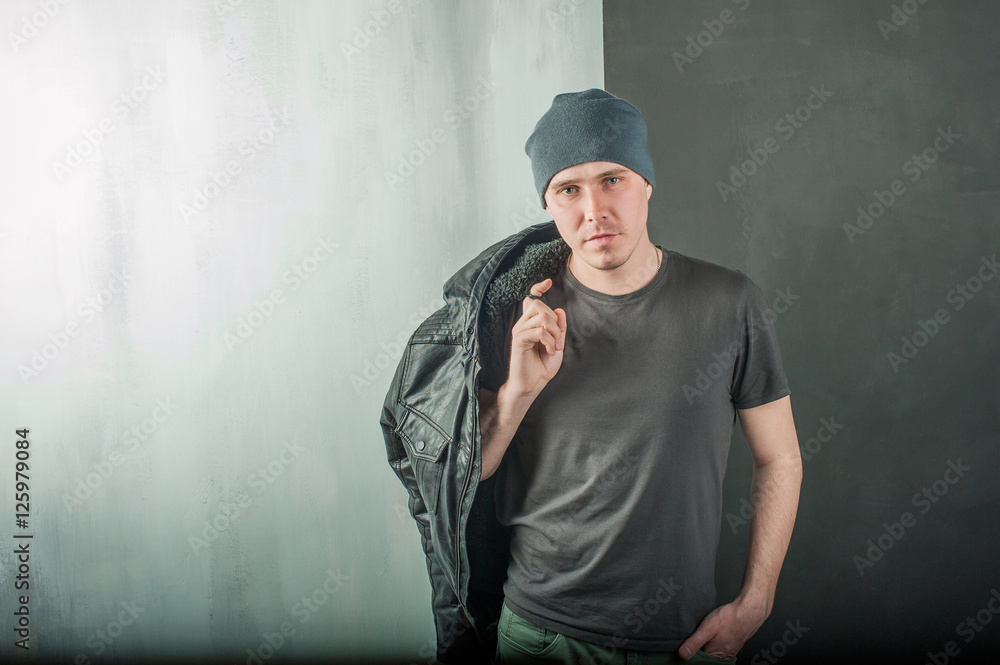 studio photography young brutal guy. man in T-shirt, jeans and a knitted cap, keeps black leather jacket, slung over his shoulder. black background painted walls and white stairs