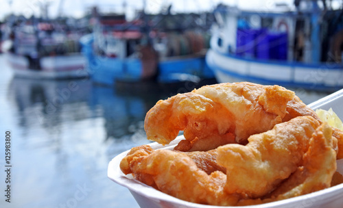 Canvas Print Battered fish served on a plate against fishing boats at sunset