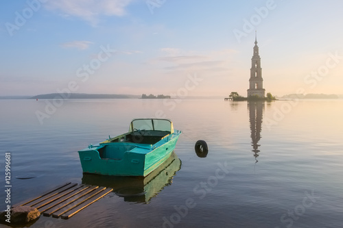 Landscape with flooded bell tower of the Cathedral of St. Nicholas in the town of Kalyazin on the Volga River in the early summer morning at sunrise with a motor boat on the shore