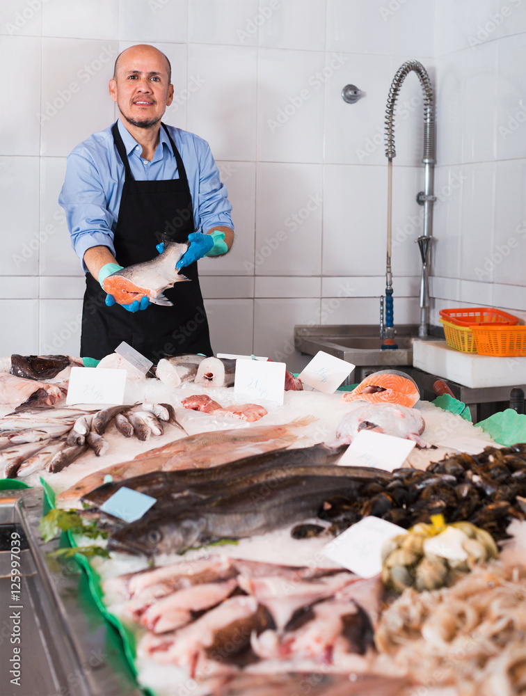 Positive cheerful mature salesman with apron offering fresh fish