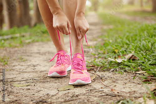 Cropped shot of young woman runner tightening running shoe laces, getting ready for jogging exercise outdoors. Female jogger lacing her pink sneakers standing on forest path before morning run © wayhome.studio 