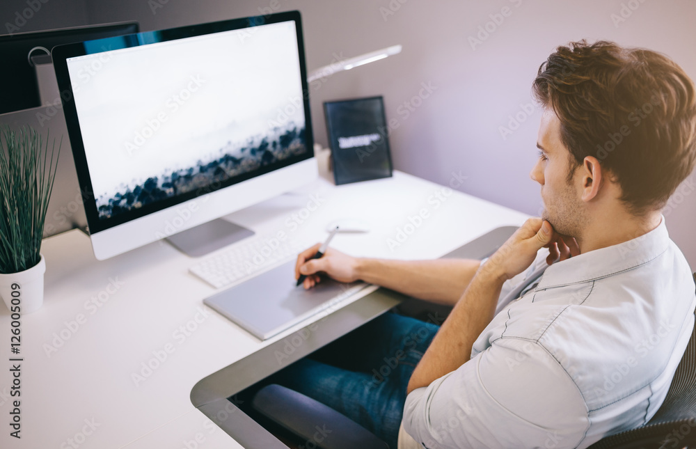 Young worker sitting in an office at the computer. Freelancer in a blue shirt. The designer sits in front of window in the workplace. Young worker sitting in an office at the computer. Freelancer in a blue shirt. The designer sits in front of window in the workplace.