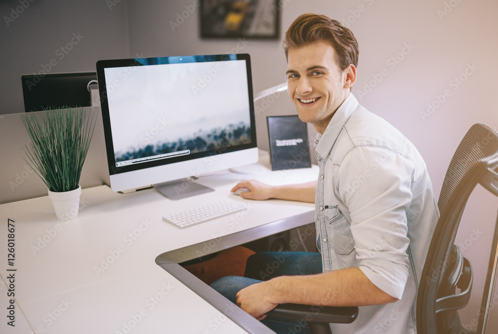 Young worker sitting in an office at the computer. Freelancer in a white shirt. The designer sits in front of window in the workplace.