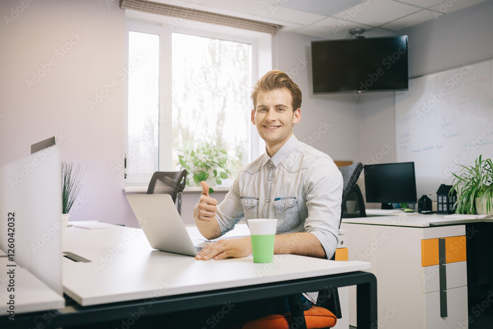 Young worker sitting in an office at the computer. Freelancer in a white shirt. The designer sits in front of window in the workplace.