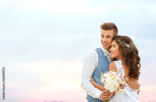 Beautiful wedding couple with bouquet of flowers against cloudy sky, close up view