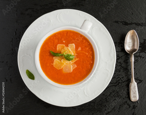 Tomato cream soup with parmesan cheese in white bowl on black stone background. Fresh basil leaves and vegetables, top view.