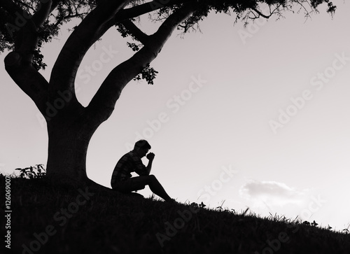 Man praying. Religion concept. Young man with his hands folded together sitting under a tree. 