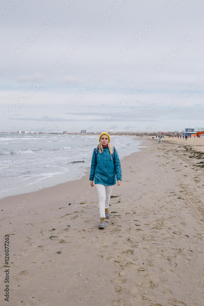 young woman traveler walking along the beach in winter. woman in a blue jacket and yellow knitted cap walking on the beach on the background stormy sea
