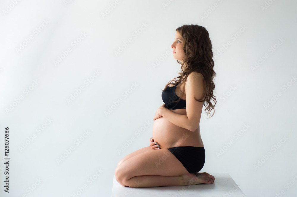 Obraz premium Sitting pregnant woman with hands on belly, white wall on background