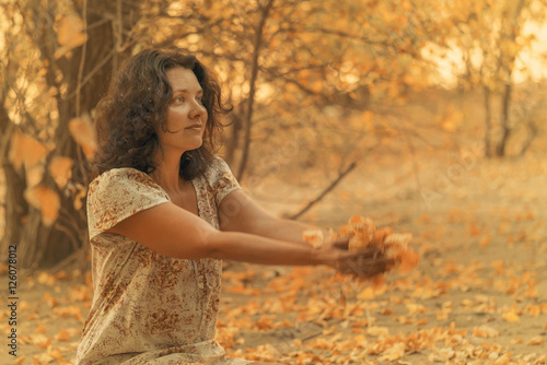 Adult brunette woman in a dress holds in hands dry yellow leaves sitting in the autumn forest