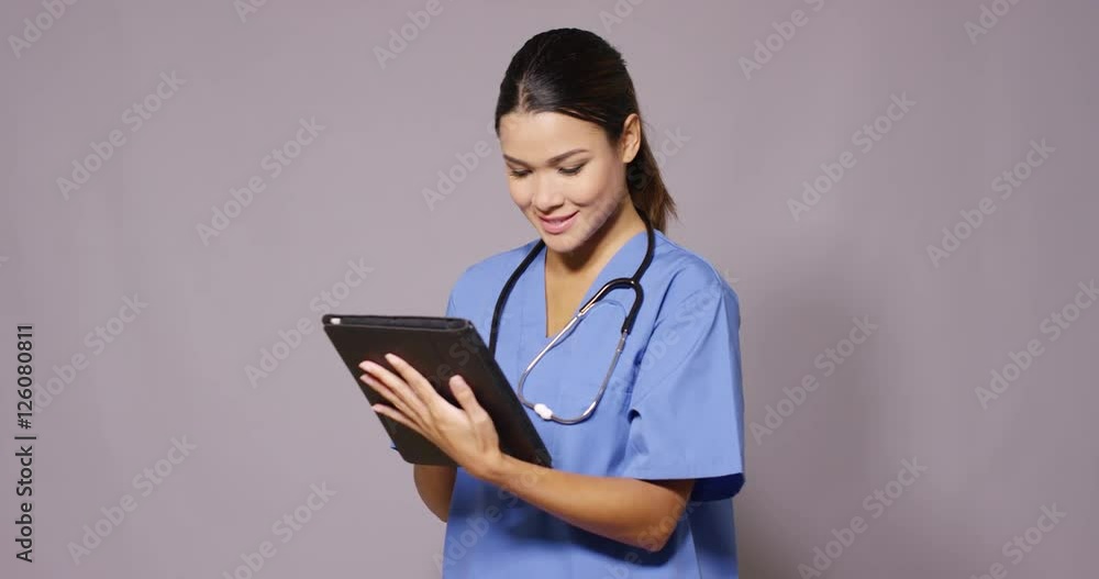 Frowning attractive young female nurse or doctor in blue surgical scrubs standing checking patient information on a tablet over grey with copy space Frowning attractive young female nurse or doctor in blue surgical scrubs standing checking patient information on a tablet over grey with copy space