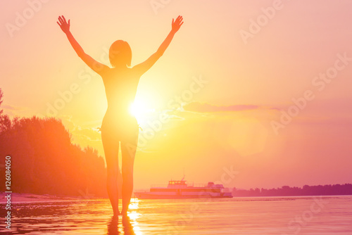 Young woman with hands raised standing in water in the sunlight, sunset or sunrise