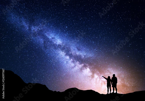 Naklejka Droga Mleczna z ludźmi na górze. Krajobraz z nocnym niebem z gwiazdami i sylwetką stojącego szczęśliwego mężczyzny i kobiety, którzy wskazując palcem w gwiaździste niebo. Droga Mleczna z podróżnikami. Piękna galaktyka