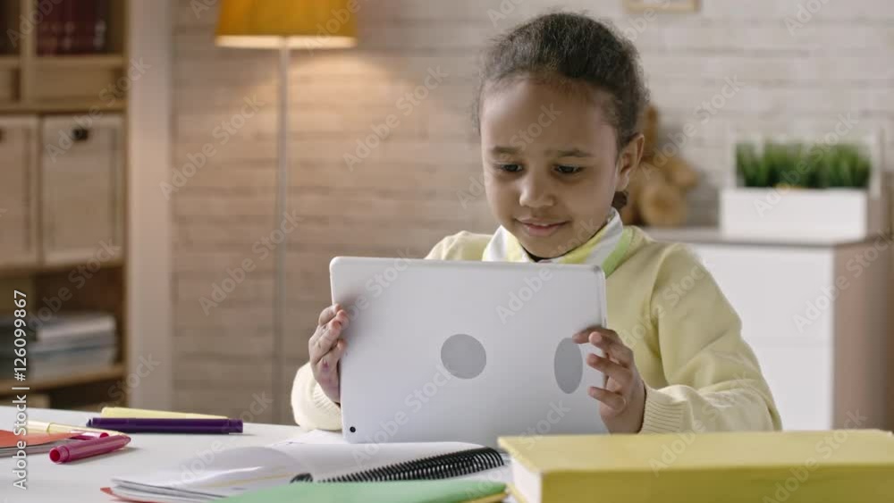 Beautiful little African girl doing homework on digital tablet at the desk Beautiful little African girl doing homework on digital tablet at the desk