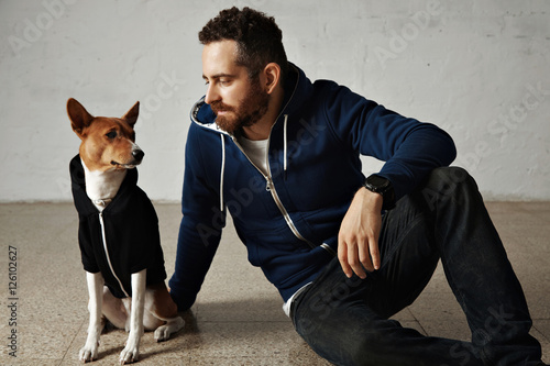 An attractive young man and his bassenji dog wearing matching hoodies sit on the floor next to white wall photo