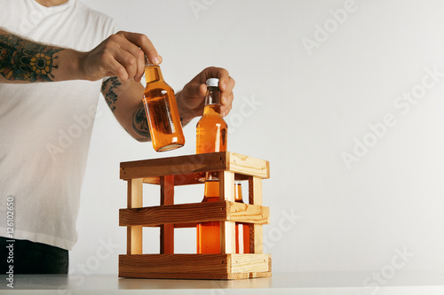 Tattooed man's hands in blank white t-shirt putting bottles with cider into a wooden crate isolated on white