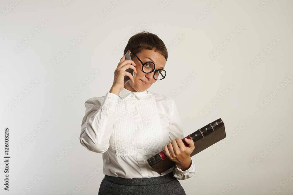 Female teacher in conservative outfit and round black glasses looks playfully into the camera while talking on the phone against white wall background
