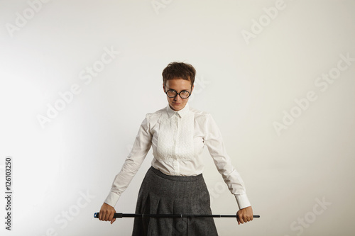 Strict looking conservatively dressed female teacher with a long black pointer against white wall background