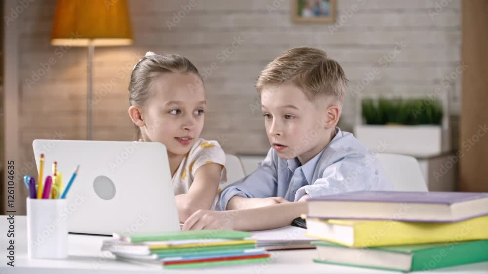 Beautiful little brother and sister sitting together at the desk at home and watching something on laptop Beautiful little brother and sister sitting together at the desk at home and watching something on laptop