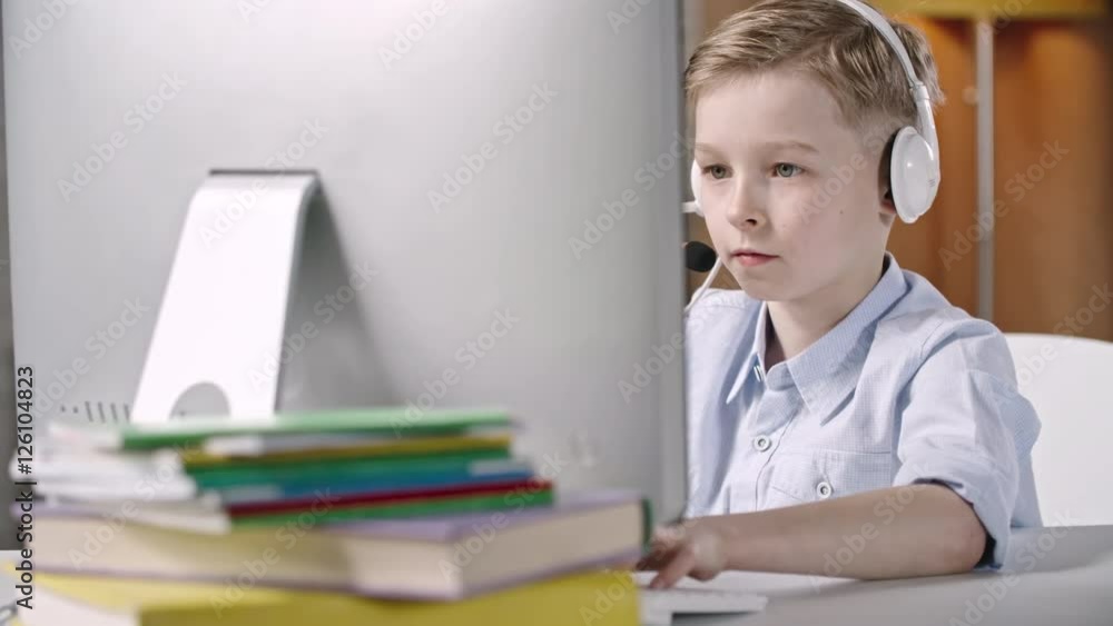 Blonde boy sitting with headset and typing something on computer at the desk Blonde boy sitting with headset and typing something on computer at the desk
