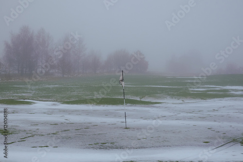 Danish golf course covered in snow, with a flag on a black and white striped pole standing in a hole, on a foggy winter day