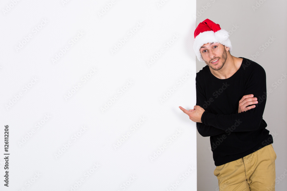 junger Mann mit Weihnachtsmann Mütze präsentiert an weißer Wand junger Mann mit Weihnachtsmann Mütze präsentiert an weißer Wand