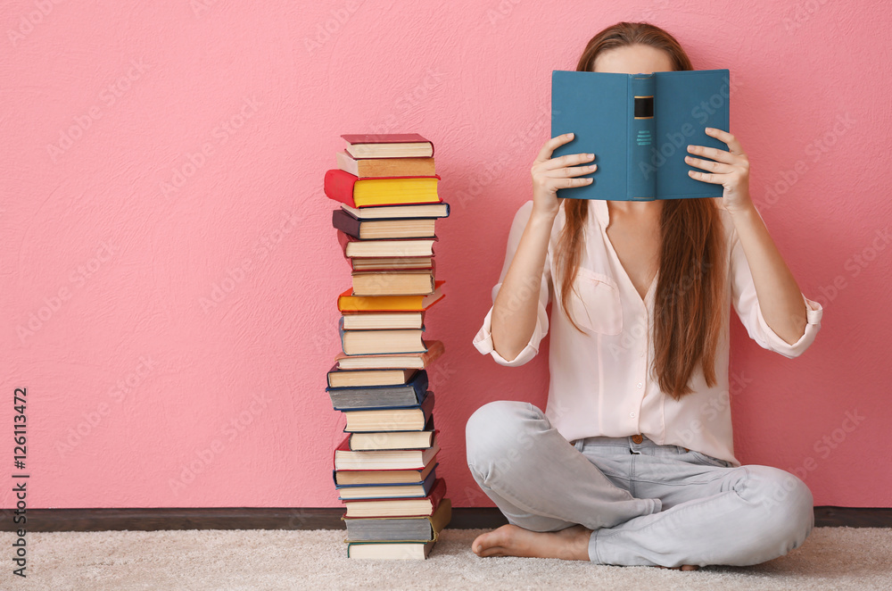 Obraz premium Woman sitting on a floor and holding book in front of face on pink background