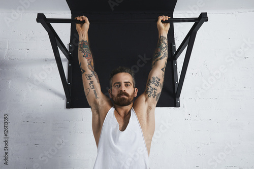 Canvas Print Brutal tattooed athletic man in white unlabeled tank t-shirt shows calisthenic moves close up of classic pullup Hanging on pull bar and looking at camera