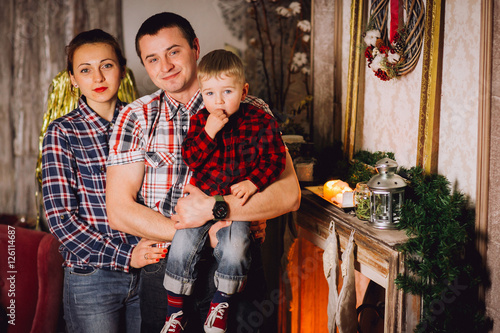 Young family is photographed for Christmas card. Mother, father and little daughter with smile look in camera. They recline on a big bed with bright pillows. Room is decorated with Christmas garlands.
