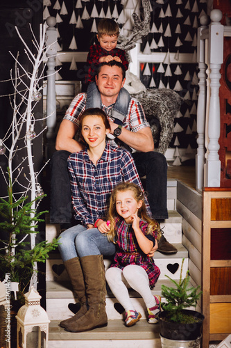 Young family is photographed for Christmas card. Mother, father and little daughter with smile look in camera. They recline on a big bed with bright pillows. Room is decorated with Christmas garlands.