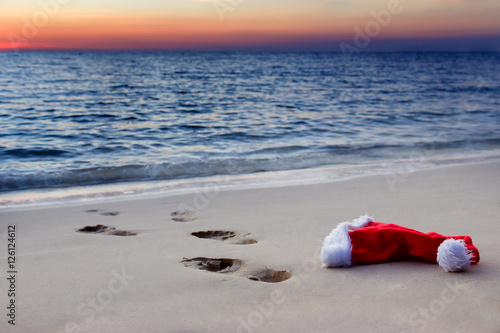 Human footprints on a sandy beach with Santa Claus hat at sunset