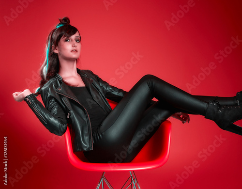 Girl in leather jacket and braided blue streaked hair lounging on red chair with bright red bkgd