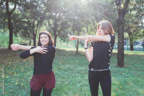 Two young beautiful caucasian women stretching outdoor in a city park in sunny day - sportive, training, wellness concept