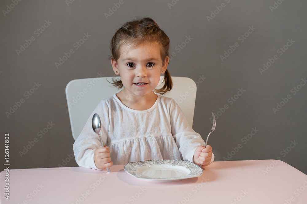 Cute little smiling girl sitting on table with plate, holding fork and spoon in hands Cute little smiling girl sitting on table with plate, holding fork and spoon in hands