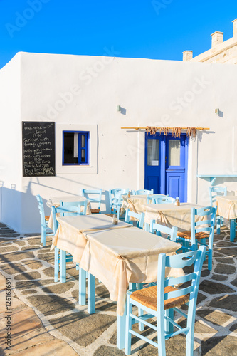 Tables with chairs in Greek taverna in Naoussa port, Paros island, Greece. Menu chalkboard on wall shows dish names with prices. © pkazmierczak