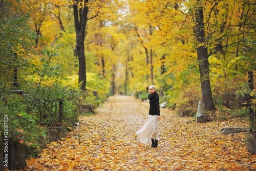 Thoughtful blonde girl in a black blouse and white skirt standing among trees in the autumn forest. © raisondtre