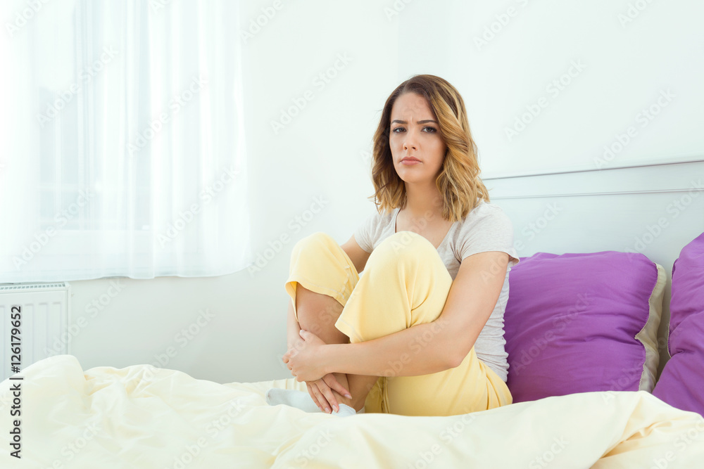 Grumpy young woman is sitting on the bed and looking at camera after waking up in the morning Grumpy young woman is sitting on the bed and looking at camera after waking up in the morning